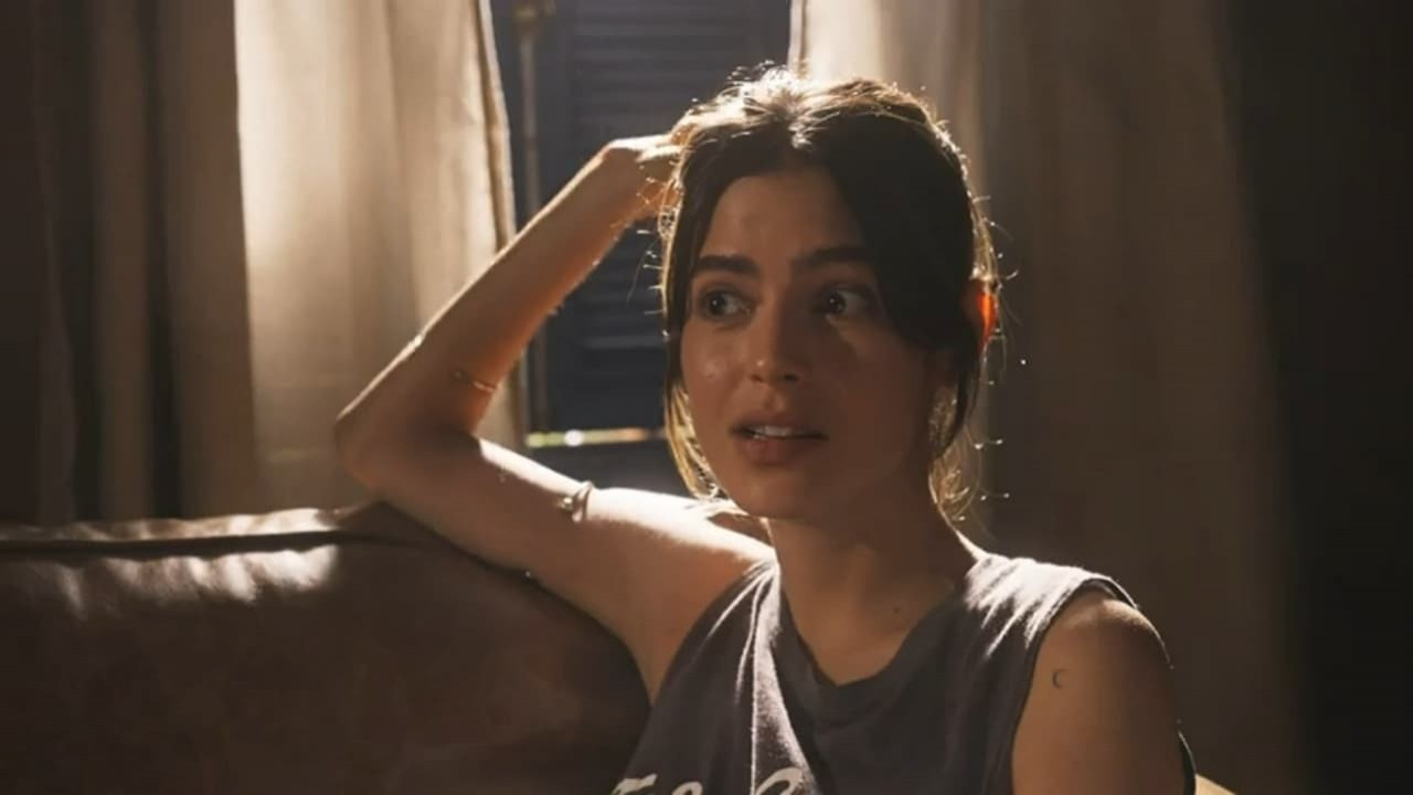 Julia Dalavia como Guta na novela Pantanal. Ela está sentada no sofá da fazenda de Tenório