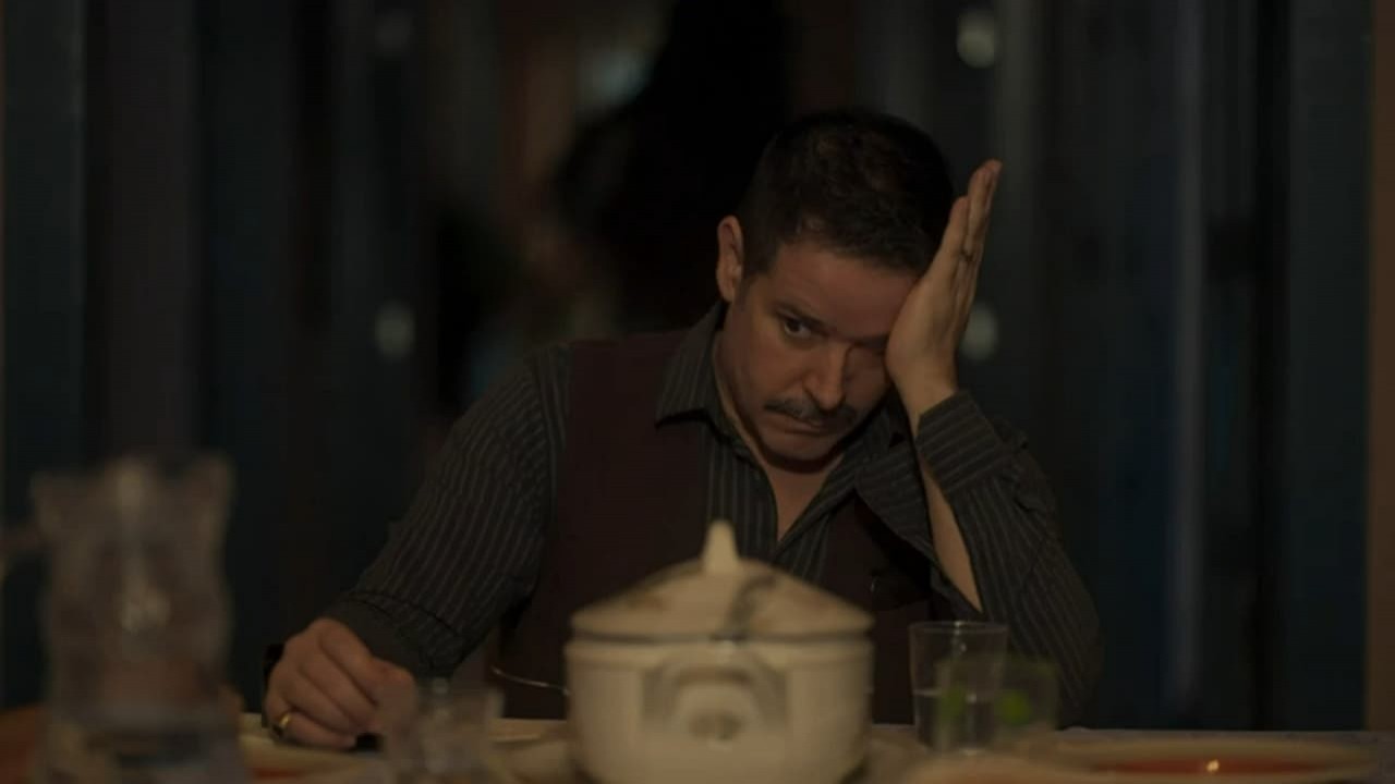 Murilo Benício em cena como Tenório na novela Pantanal. Ele está sentado e com o braço em cima da mesa da cozinha