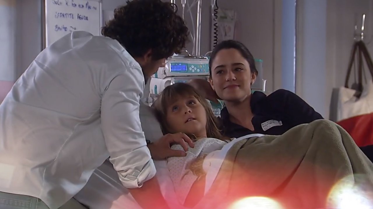 Júlia deitada na cama do hospital com Ana falando com Rodrigo que está de pé