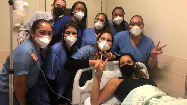 Ivy Moraes, em leito hospitalar, posa com técnicas de enfermagem do hospital