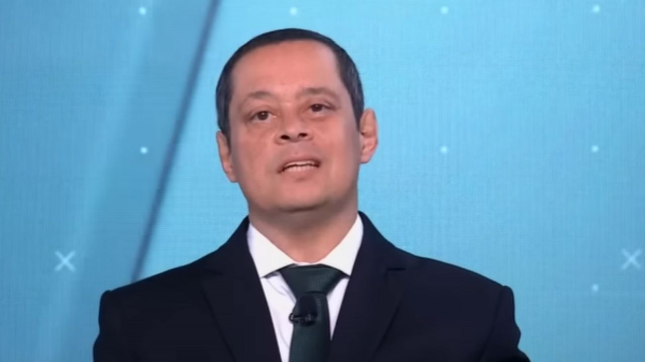 Jorge Serrão falando em fundo azul, de terno e gravata