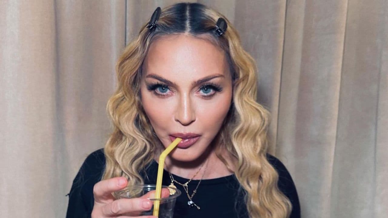 Madonna bebendo algo em copo com canudo amarelo, olhando para a câmera, de camiseta preta