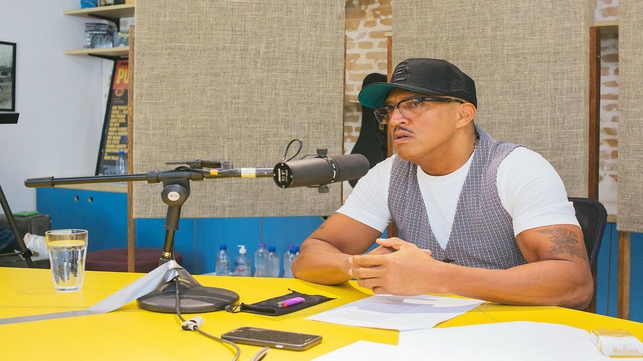 Mano Brown  sentado em uma cadeira com um microfone na sua frente na mesa do podcast Mano a Mano do Spotify