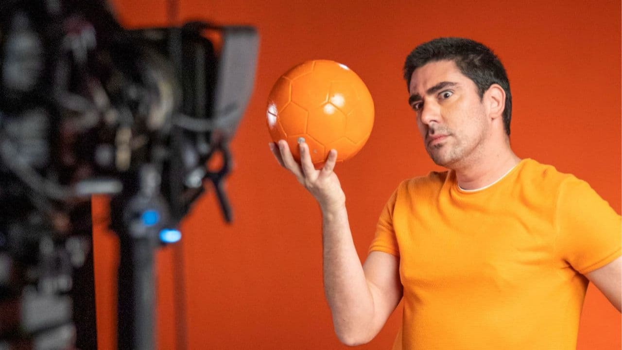 Marcelo Adnet posando para câmera, com camiseta laranja e bola na mão
