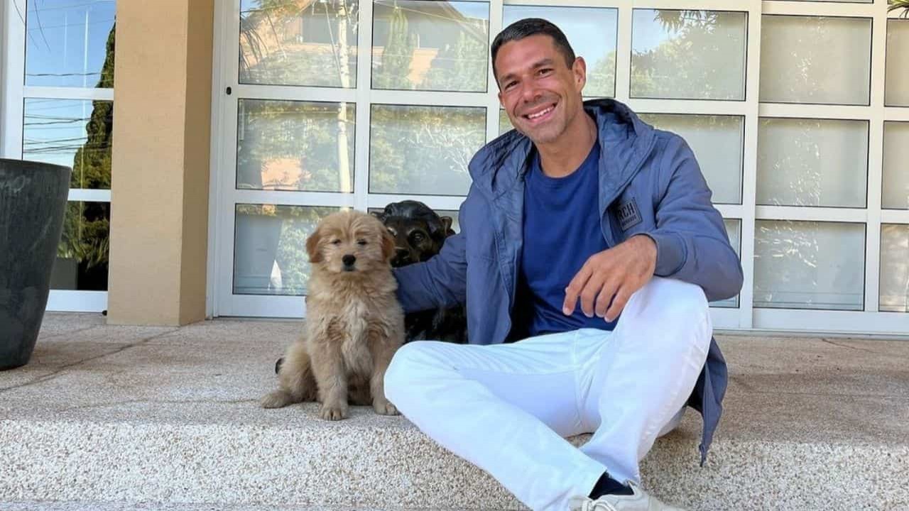 Marcus Buaiz de calça clara, blusa e casaco azuis, sorrindo para foto ao lado de cachorrinho