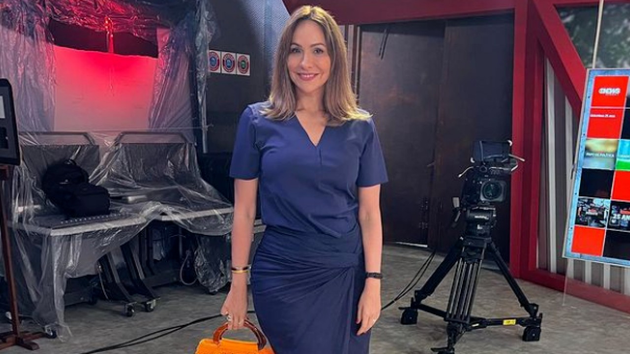 Maria Beltrão com vestido azul e bolsa laranja pousando para foto dentro de estúdio da GloboNews
