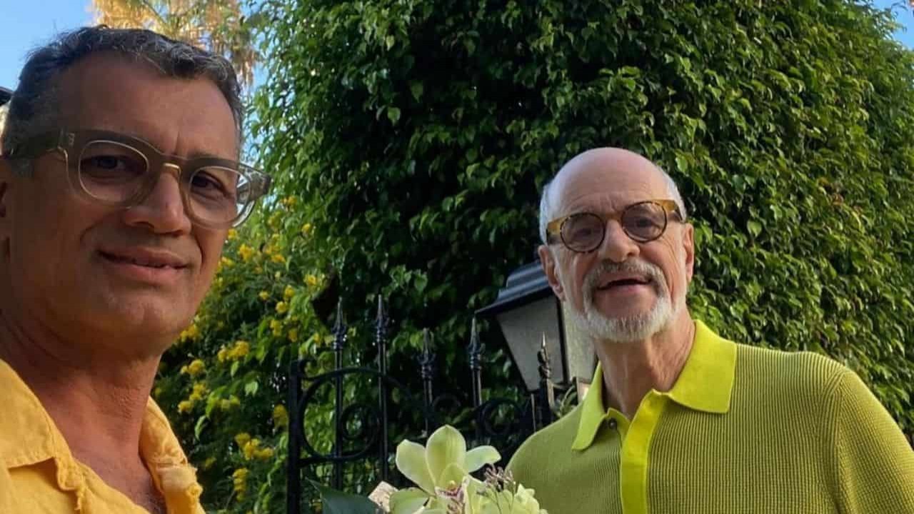 Marcos Paiva e Marcos Caruso posando para foto perto de flores, com roupas amarelas e óculos de grau