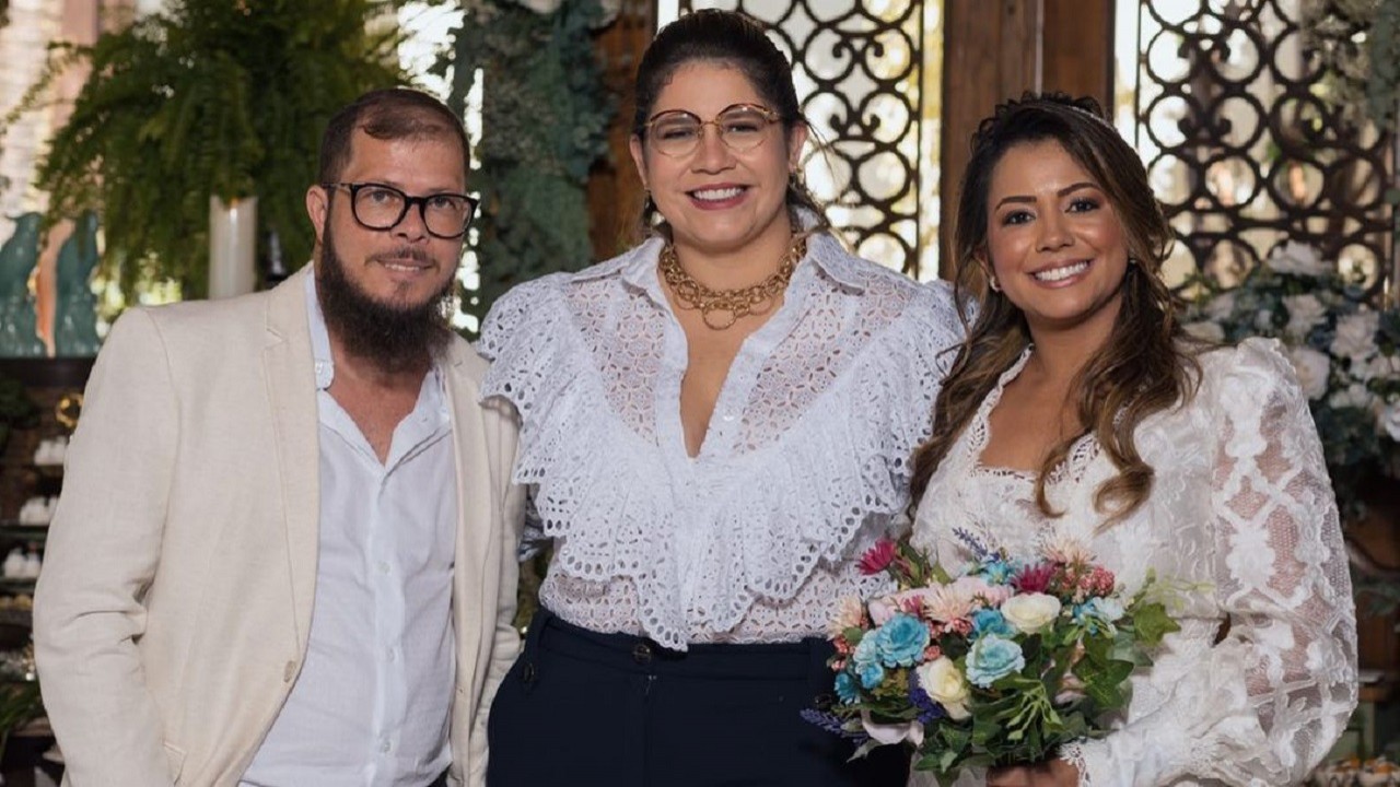 Marília Mendonça no meio, sorridente ao lado do seu tio, Abicieli Silveira Dias Filho e a mulher dele, Nayara Moura, vestida de noiva, segurando um buquê 