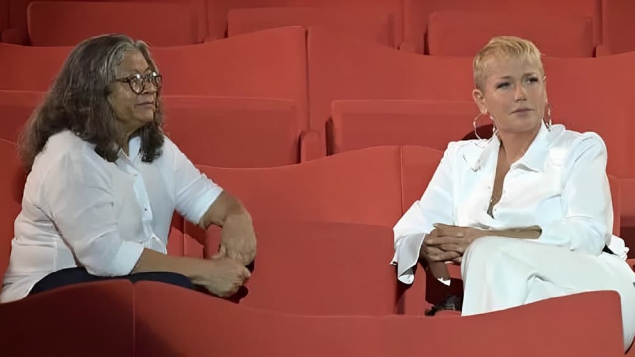 Marlene Mattos e Xuxa Meneghel com expressões sérias, sentadas em poltronas vermelhas