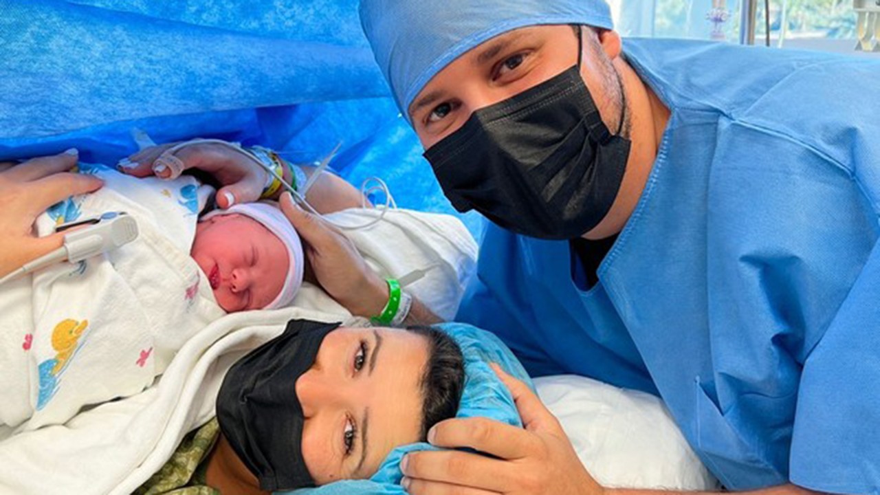 Mulher Dony de Nuccio em leito hospitalar segurando filho recém-nascido, com jornalista tocando mulher e bebê. Todos posados.