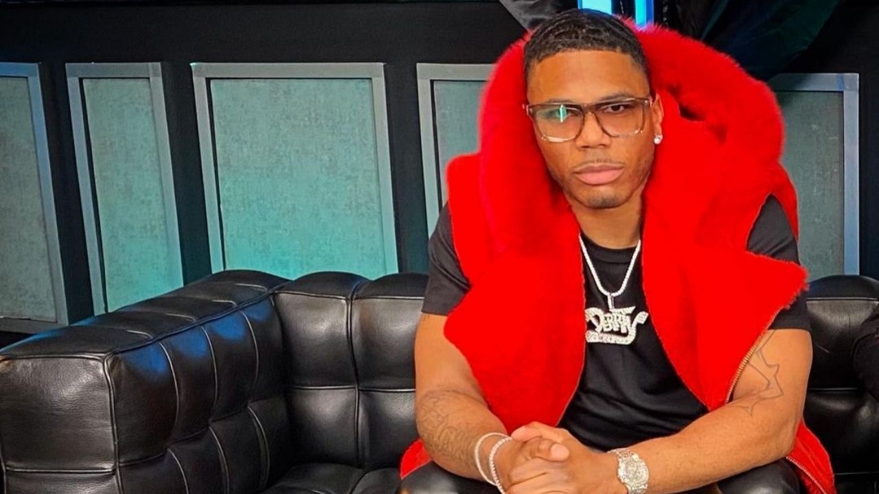 Nelly sentado em sofá preto com expressão série e mãos dadas, vestindo roupa preta e colete vermelho
