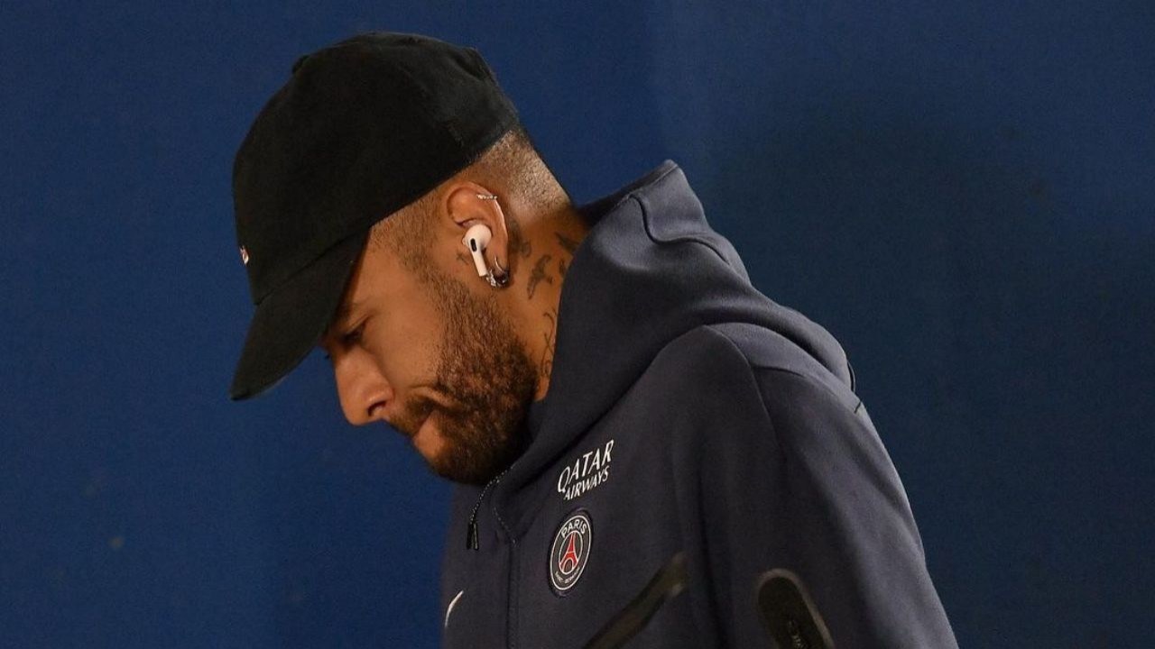 Neymar de moletom azul marinho e boné preto, de perfil com a cabeça baixa e fones de ouvido brancos