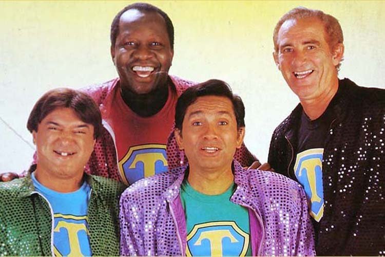 Foto dos Trapalhões Zacarias, Mussum, Dedé e Didi sorridentes usando jaquetas coloridas e camiseta com a letra T