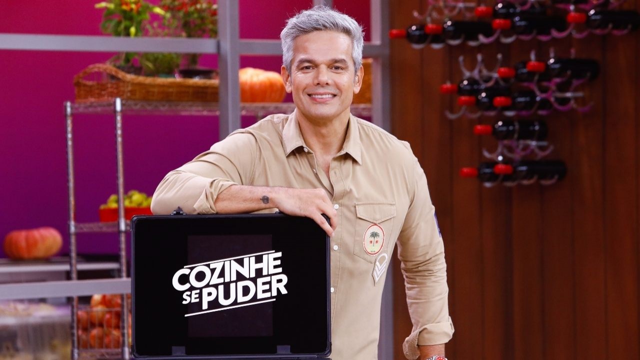Otaviano Costa de camisa bege, sorrindo e posando para foto com braço direito em cima de maleta preta do Cozinhe se Puder