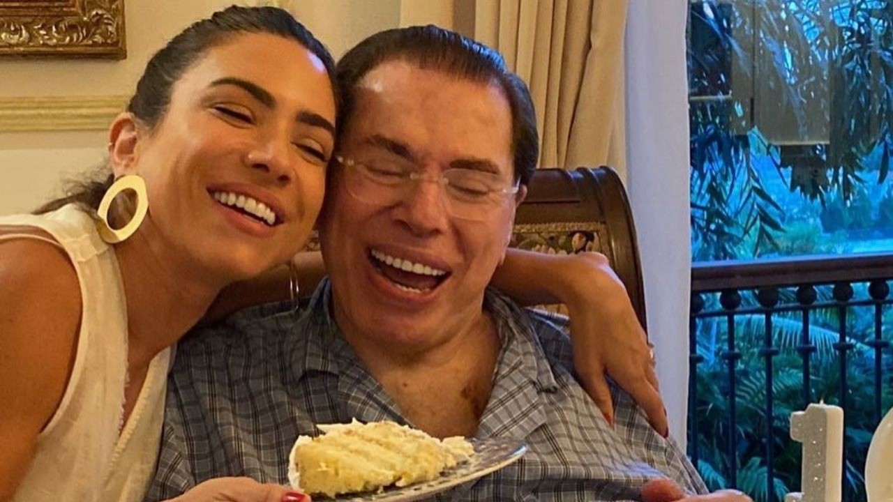Patrícia Abravanel com um pedaço de bolo na mão, abraçando Silvio Santos e sorrindo