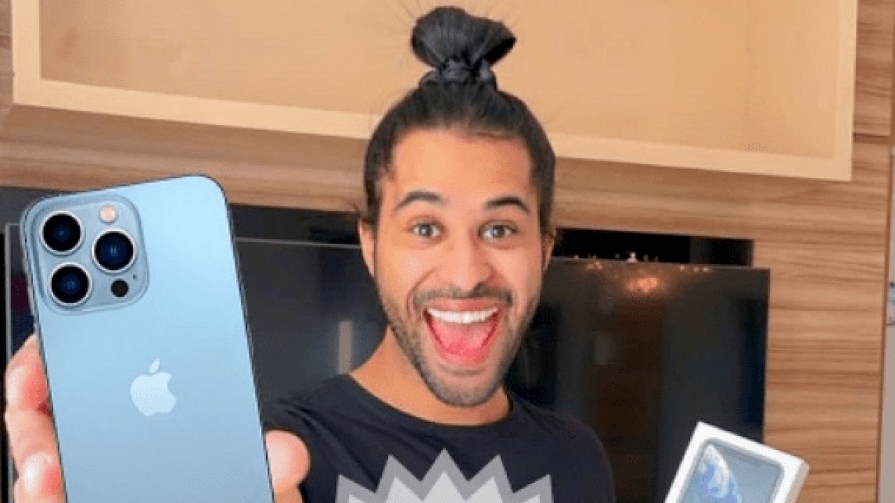 Luciano Estevan sorrindo, segurando aparelho de celular e coque nos cabelos