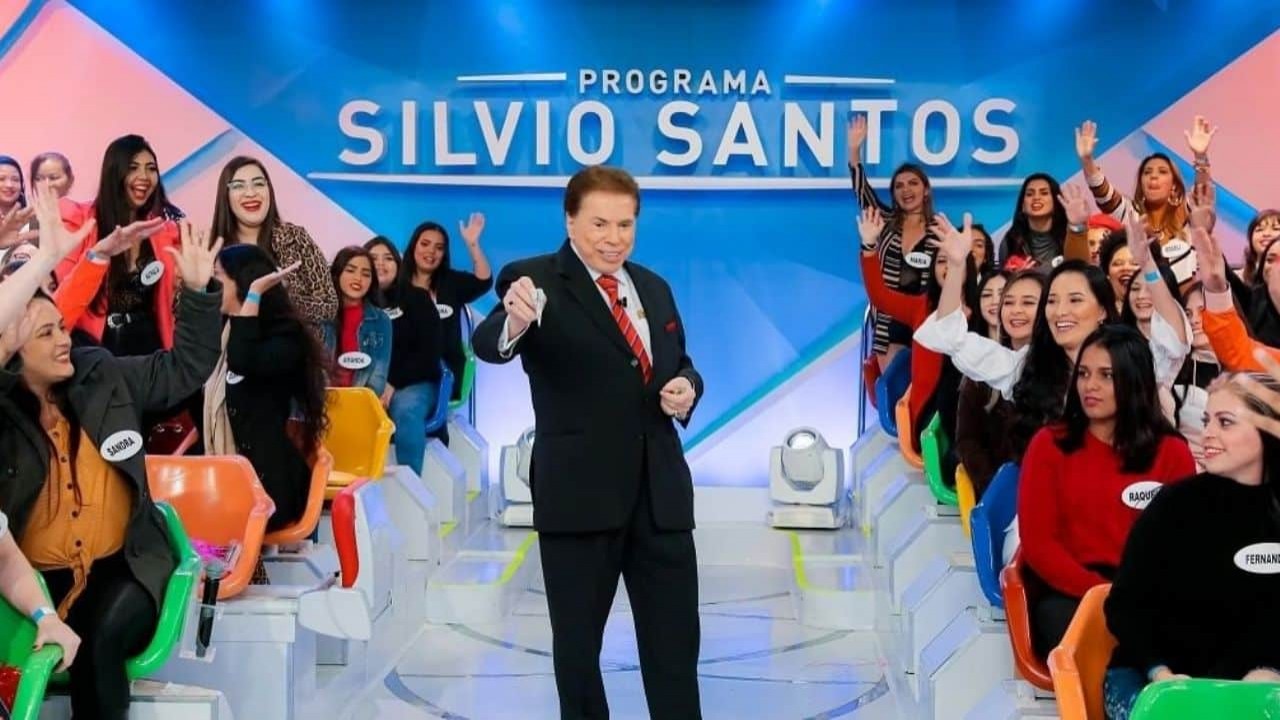 Silvio Santos perto de plateia de seu programa, vestindo terno preto e com o braço estendido