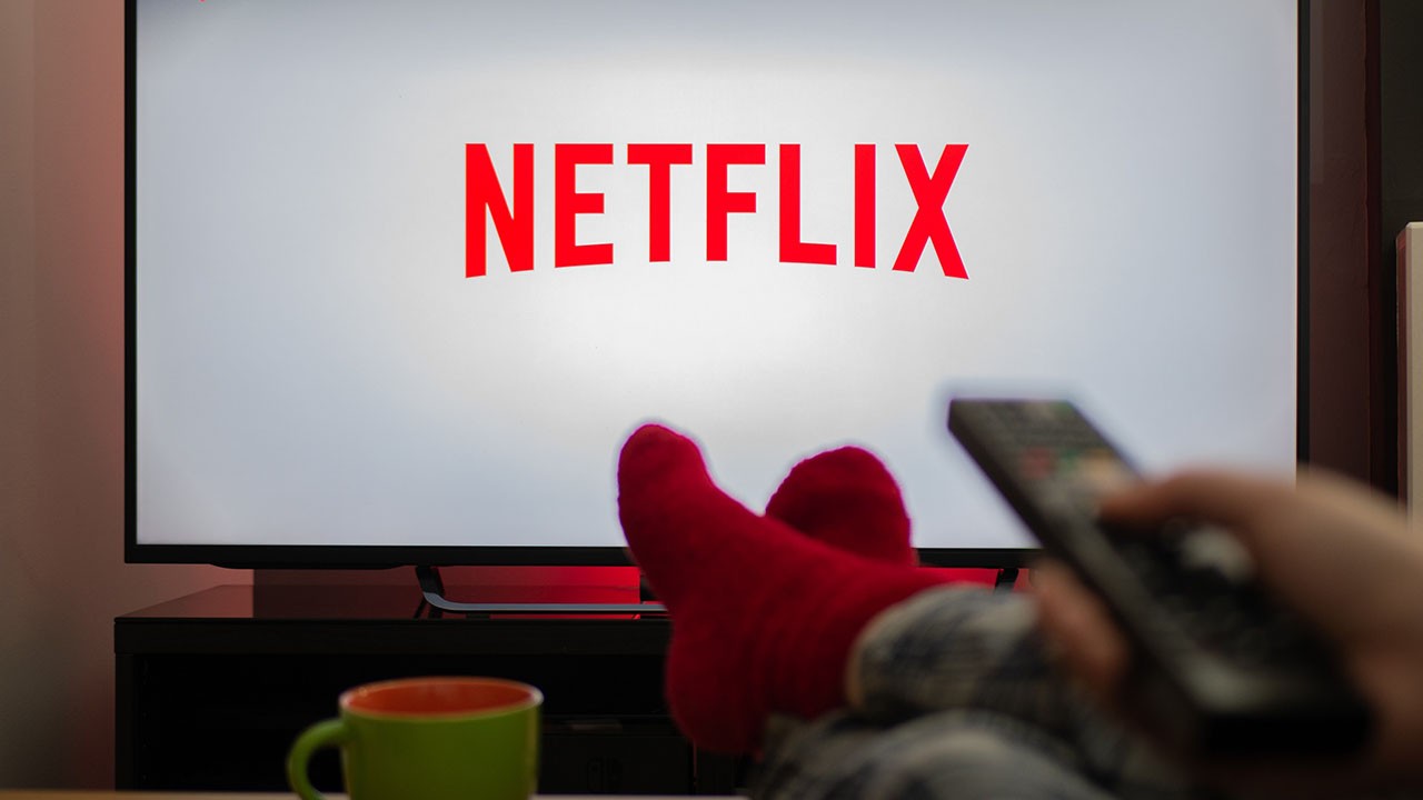 Logo na Netflix aparecendo na tela da televisão, com pessoa com pés com meia sobre a mesa, ao lado de uma caneca, segurando um controle remoto