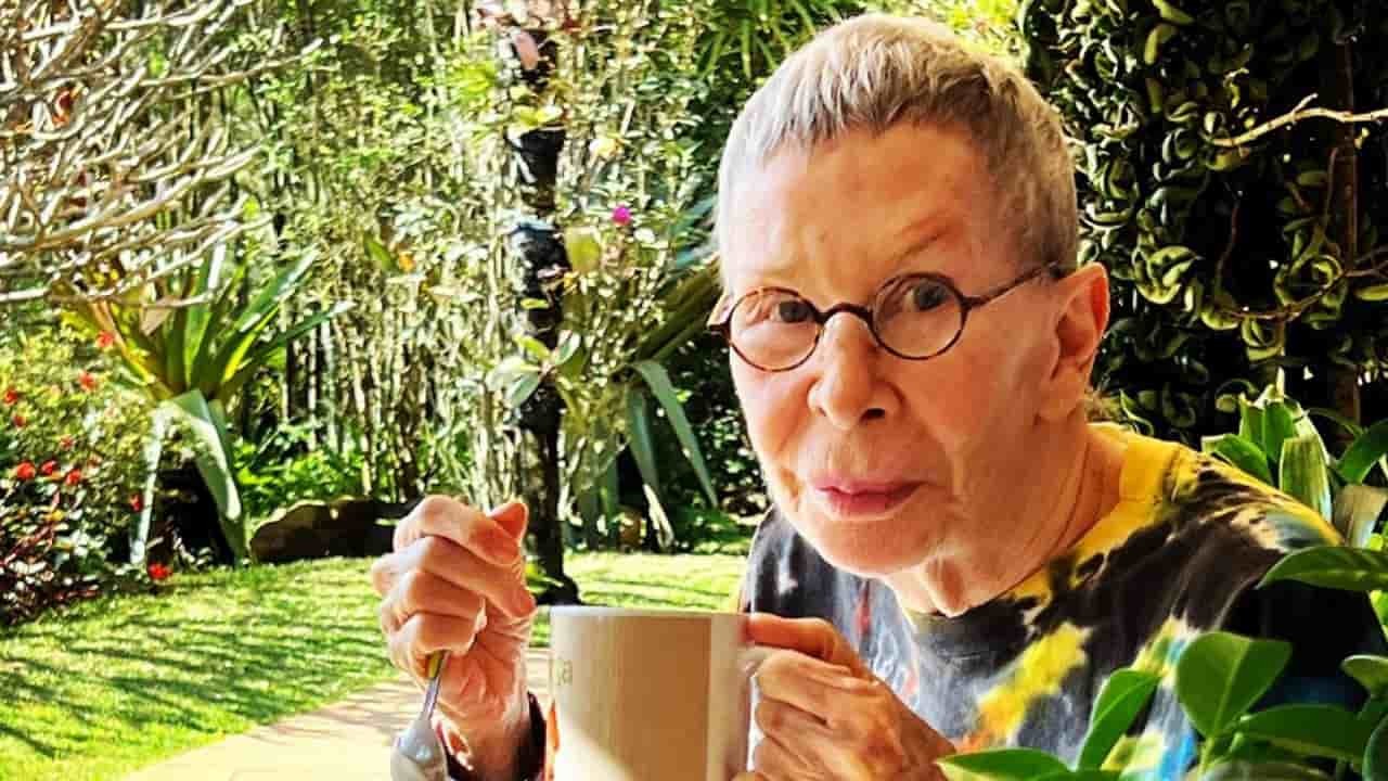 Rita Lee surge de cabelos curtos e grisalhos cercada por árvores em seu sítio no interior de São Paulo. A cantora de 74 anos segura uma caneca e uma colher