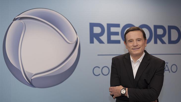 Roberto Cabrini posa sorrindo tendo ao fundo o logotipo da Record