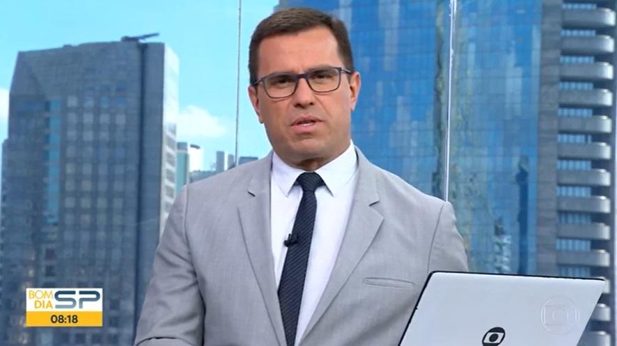 Rodrigo Bocardi de terno cinza, gravata azul escura com um computador com logo da Globo
