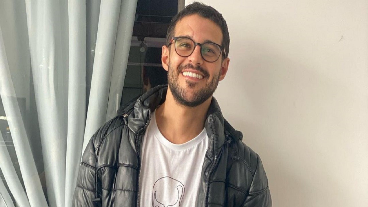 Rodrigo Mussi, sorridente, de óculos de grau, jaqueta preta e barba crescida
