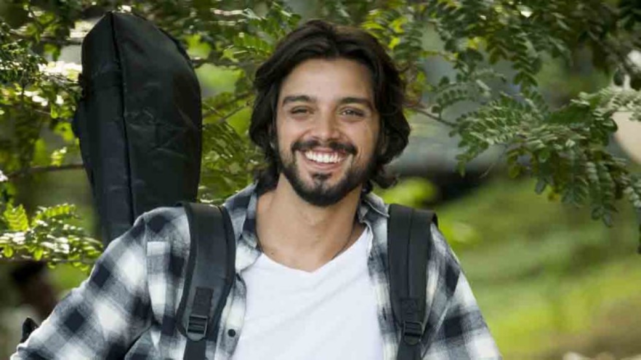 Rodrigo Simas de camisa xadrez e mochila nas costas sorrindo