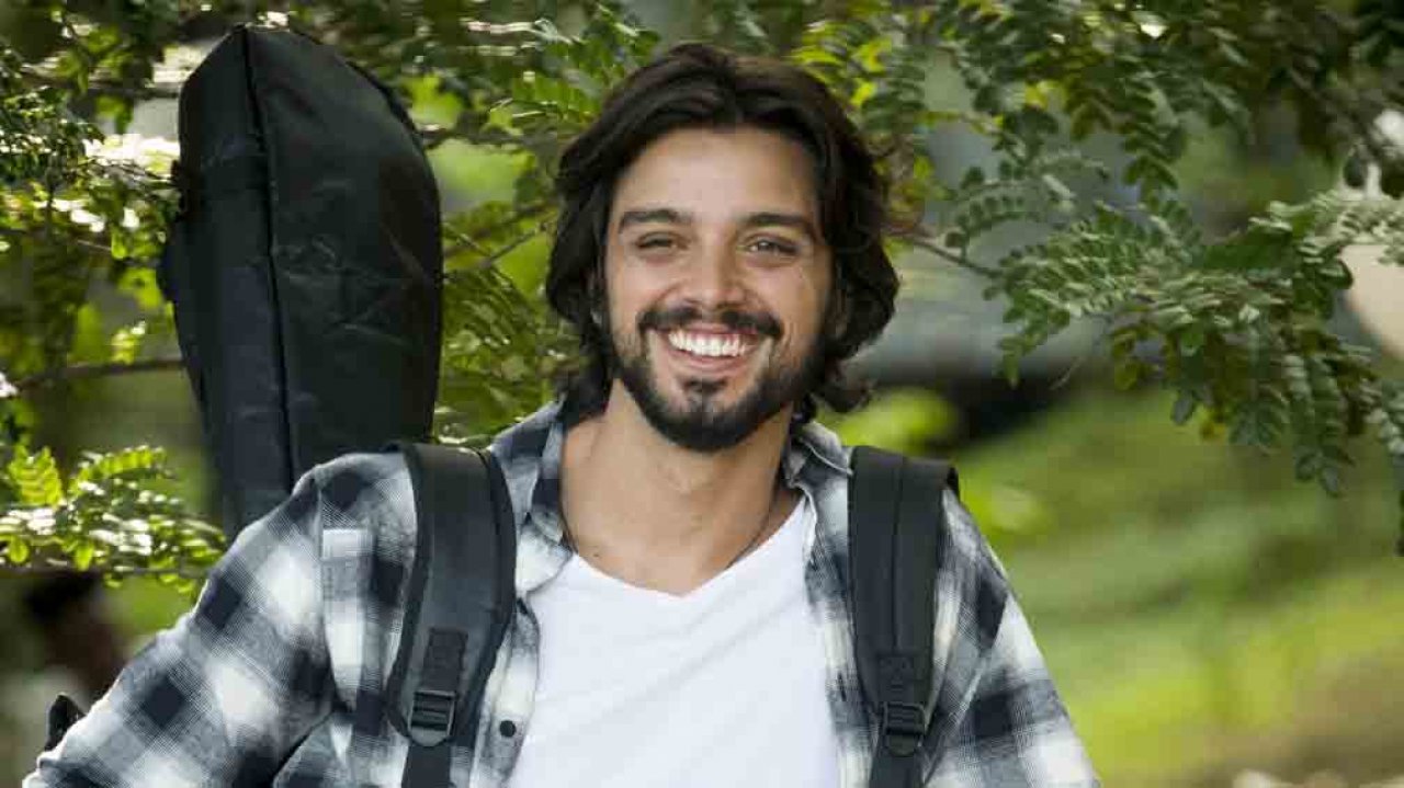 Rodrigo Simas sorrindo com violão nas costas de camisa xadrez