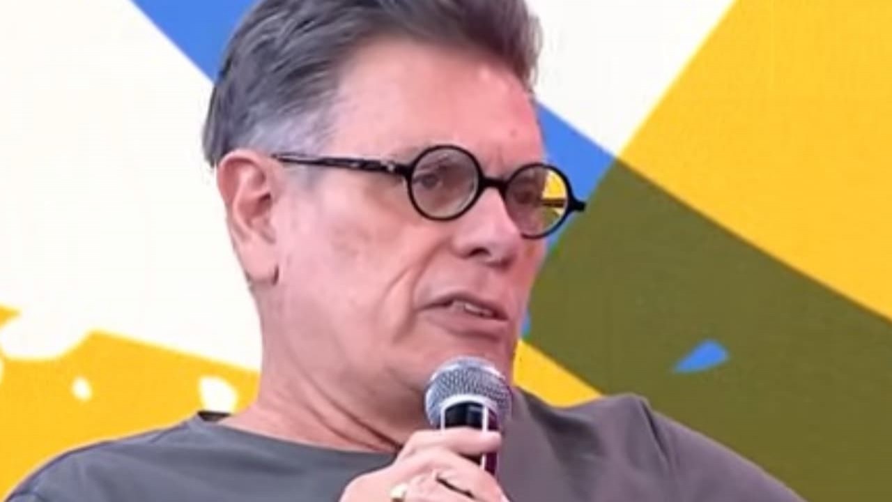 Roger Moreira em entrevista ao Pânico, de camiseta cinza e óculos, falando em microfone, sério, sem olhar para a câmera