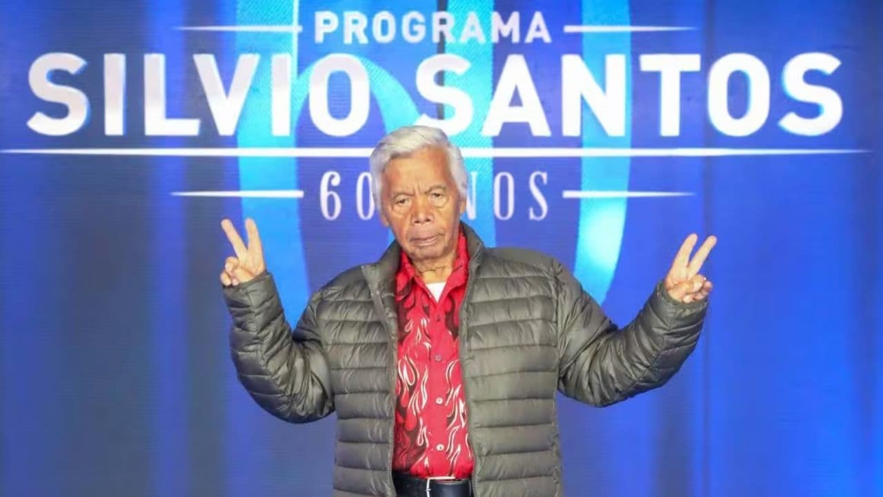Roque com expressão séria e fazendo sinal de "paz e amor" no estúdio do Programa Silvio Santos