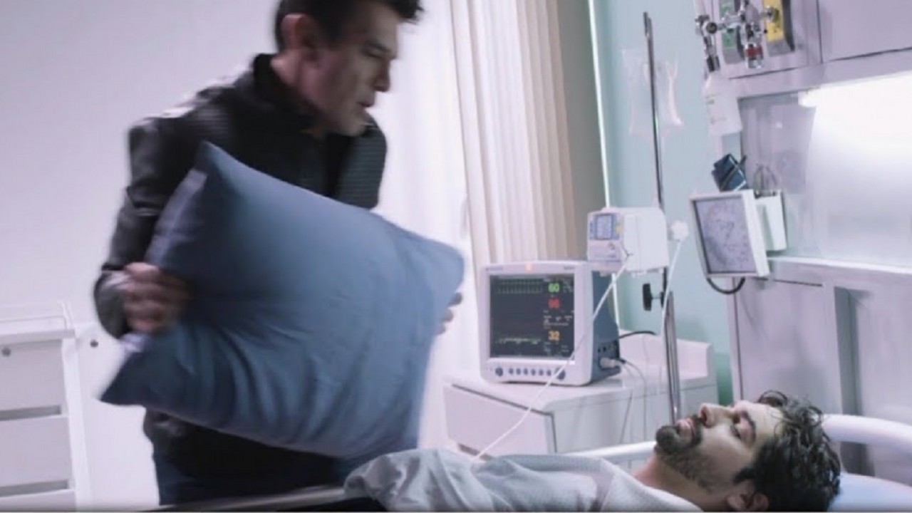 Cena de Te Dou a Vida com Ernesto segurando um travesseiro, em pé, à beira da cama de Jaime, que dorme, no quarto de hospital