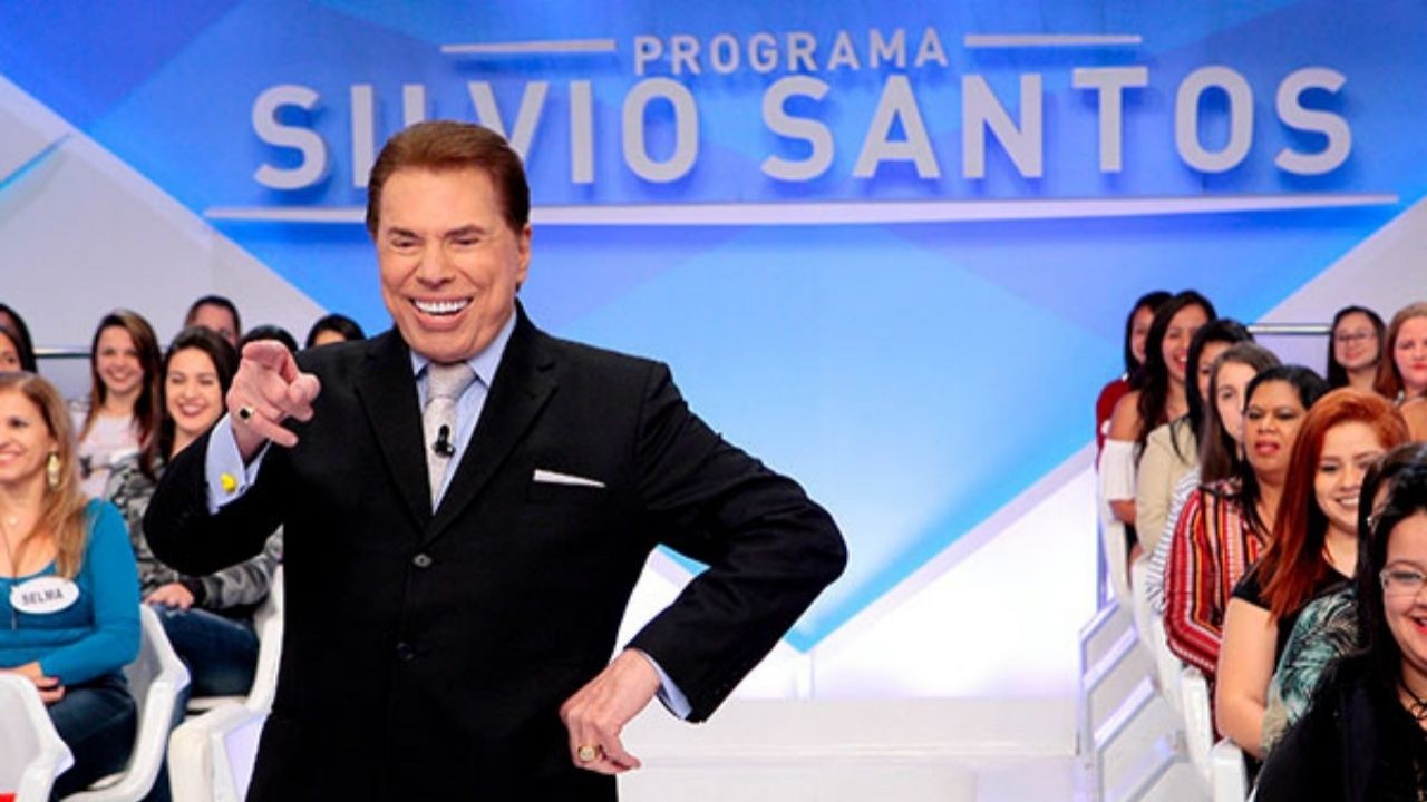 Silvio Santos rindo e apontando perto de plateia no cenário de seu programa no SBT