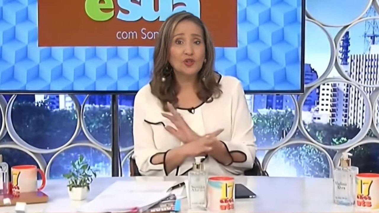 Sonia Abrão falando e gesticulando, de roupa de mangas compridas branca, no cenário de seu programa