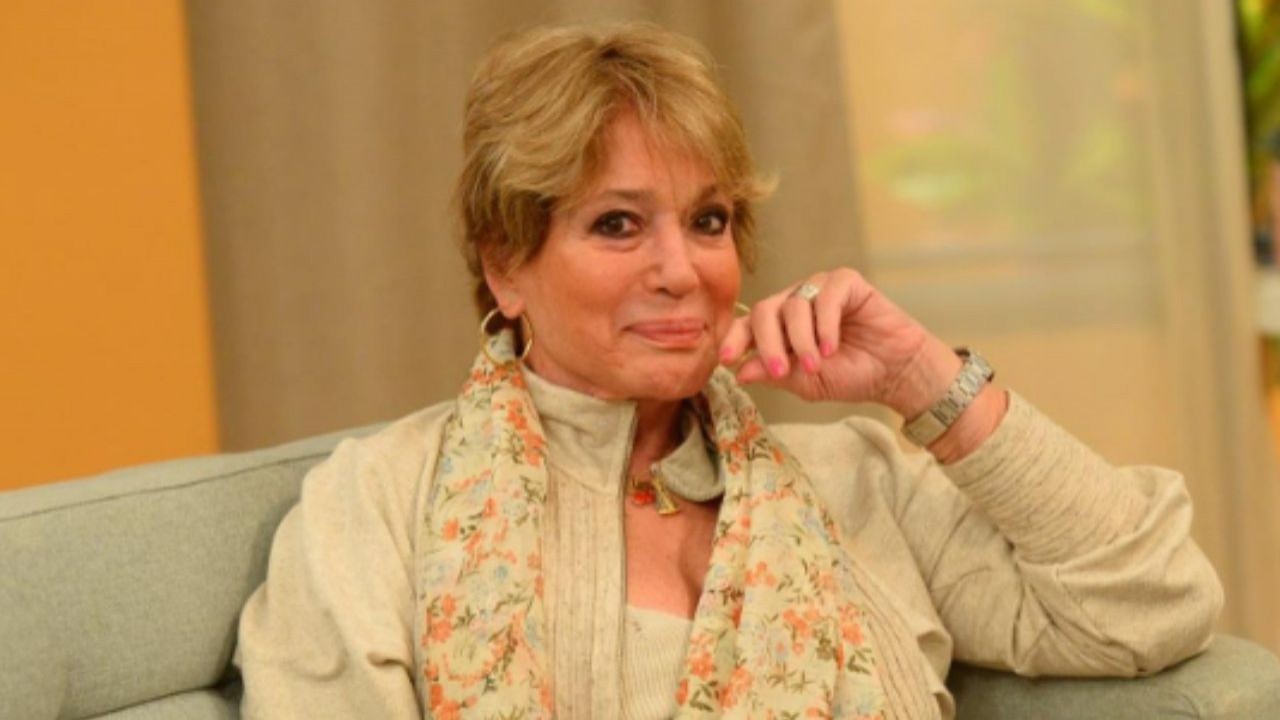 Susana Vieira de roupa bege e lenço colorido, posando para foto sentada em sofá