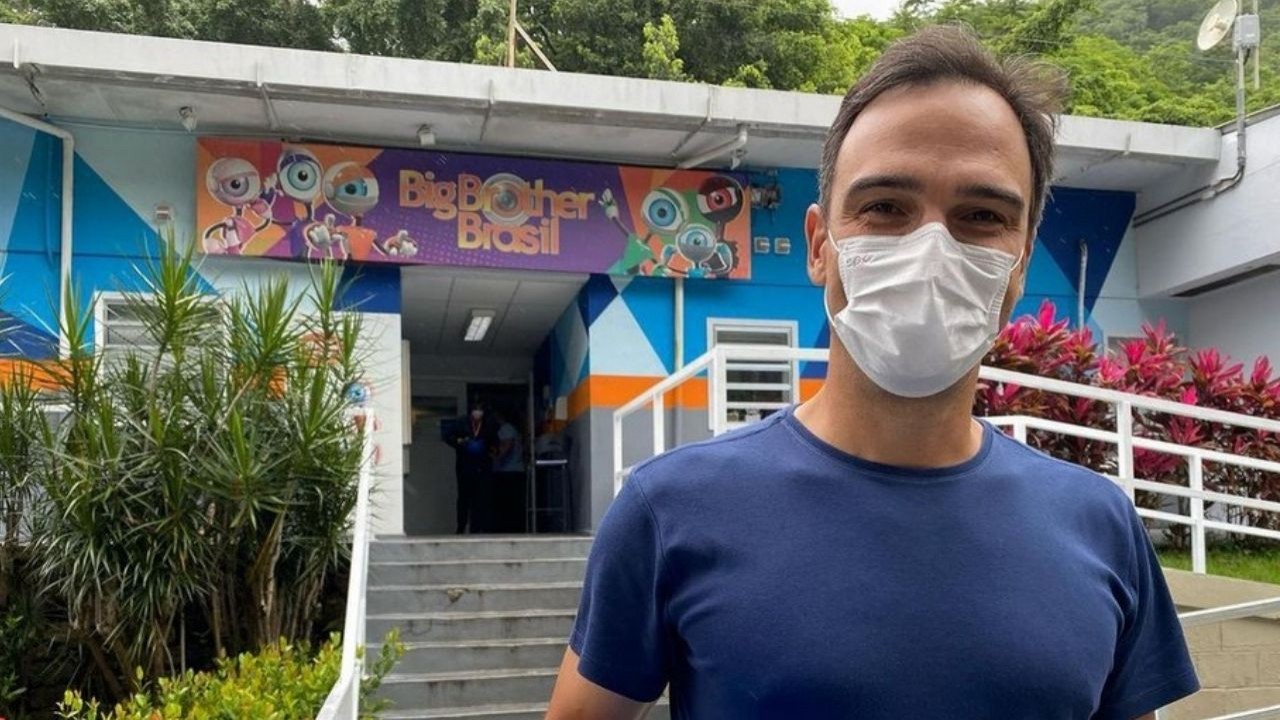 Tadeu Schmidt posando para foto em obra da casa do Big Brother Brasil, de máscara e camiseta azul