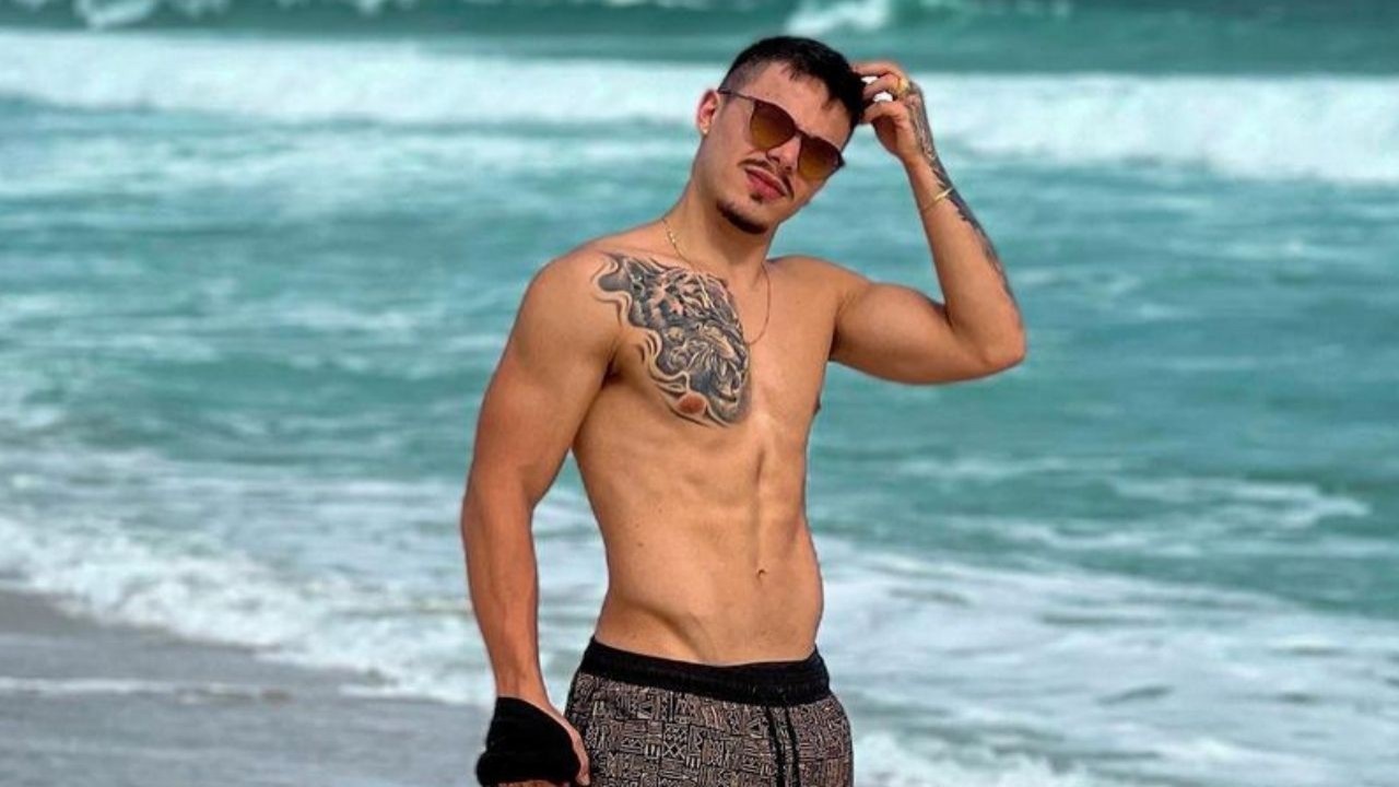 Thomaz Costa de bermuda, sem camisa, usando óculos escuros e passando a mão no cabelo, na praia
