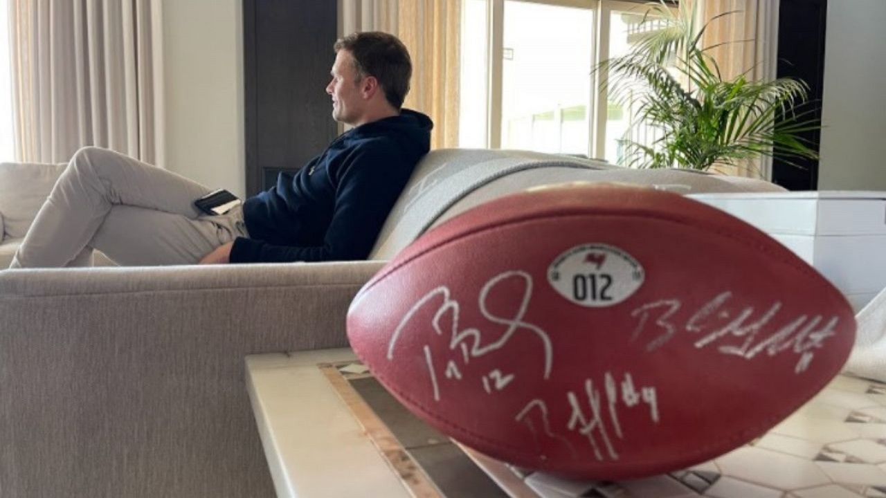 Tom Brady sentado no sofá em segundo plano com uma bola de futebol americano em primeiro plano