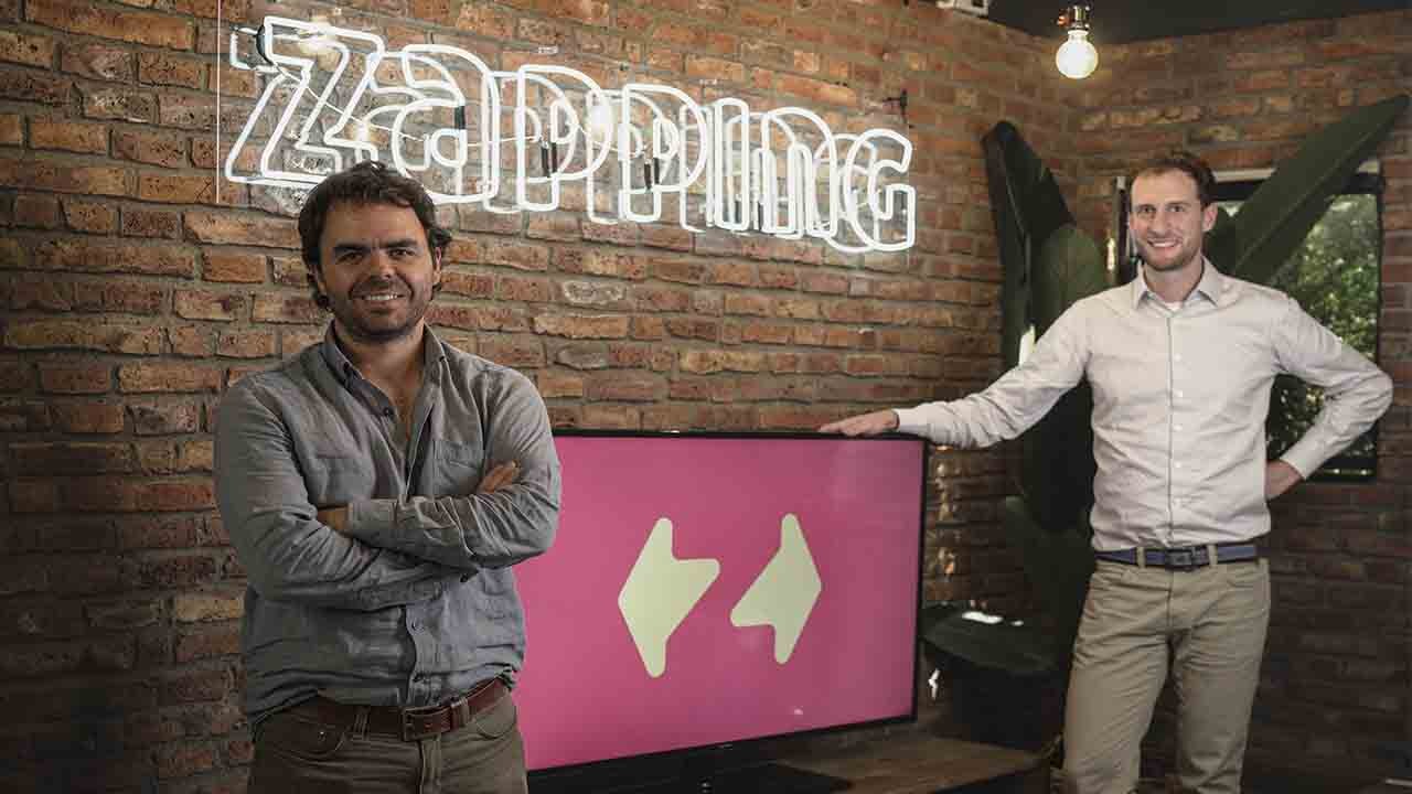 Sócios da Zapping posando ao lado do logotipo da empresa