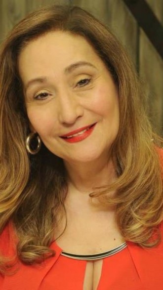 Sonia Abrão sorrindo, vestindo blusa coral, de cabelos soltos e brincos de argolas douradas